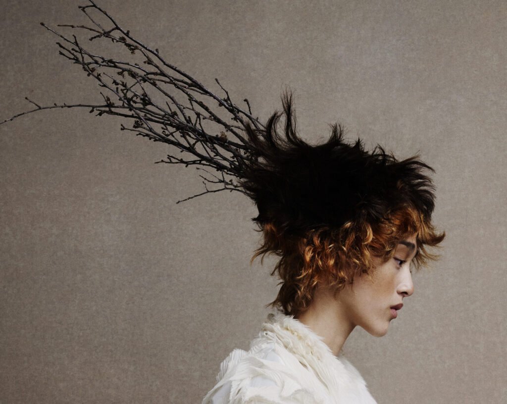 Profile of a model with a short, spiky textured crop shifting from black roots to copper curls, a bare branch woven into the hair, worn with a textured ivory garment
