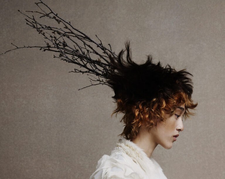Profile of a model with a short, spiky textured crop shifting from black roots to copper curls, a bare branch woven into the hair, worn with a textured ivory garment