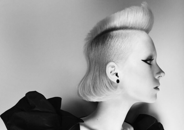 Side profile of a sleek white-blonde bob with a raised textured quiff at the crown and smooth jaw-length sides, styled with dramatic winged eyeliner