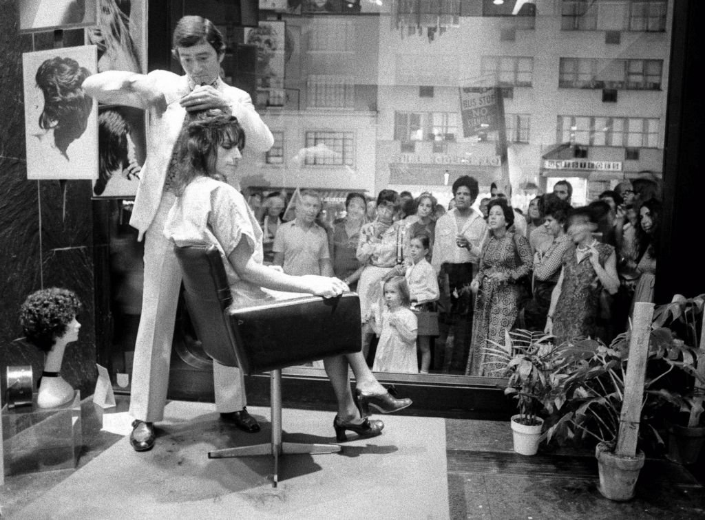 Black and white photograph of a hairstylist cutting a seated client's hair beside a salon window while a crowd of passers-by watches from the street outside.