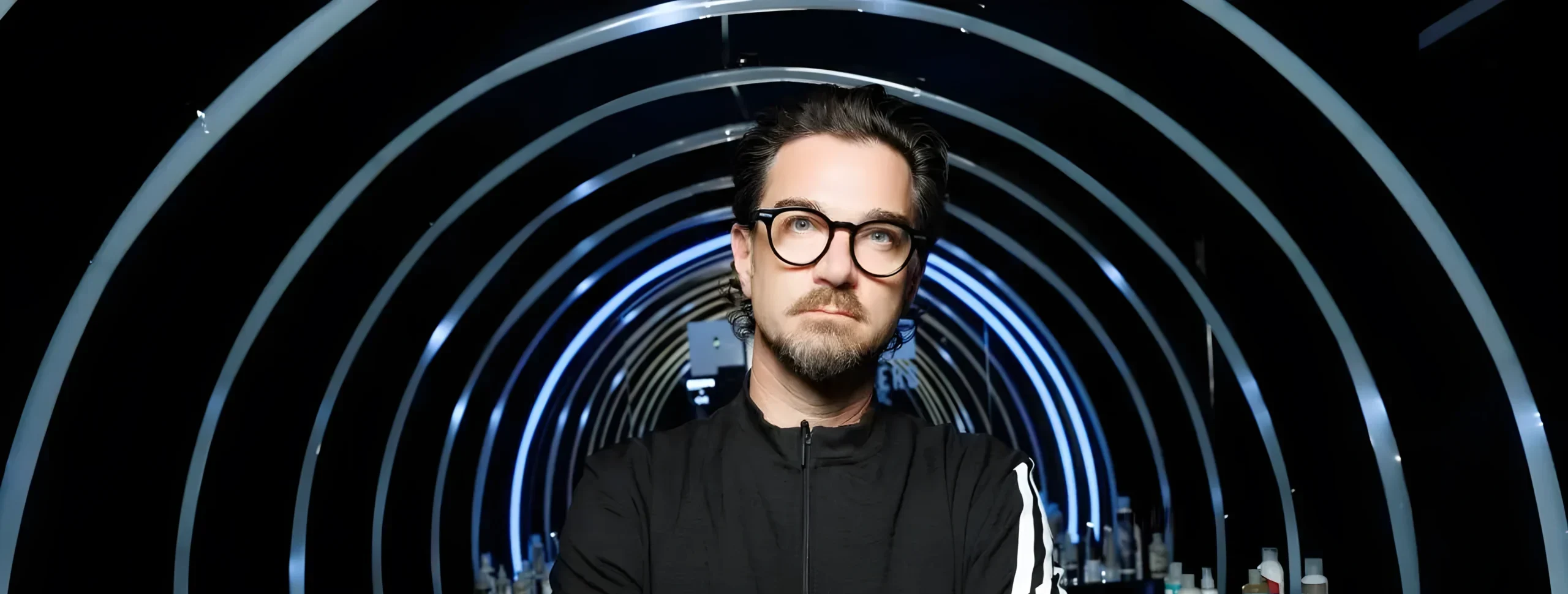 Adam Federico with swept-back dark hair, round dark glasses and a goatee, arms crossed in a black track jacket, standing in a tunnel of illuminated rings lined with haircare product displays