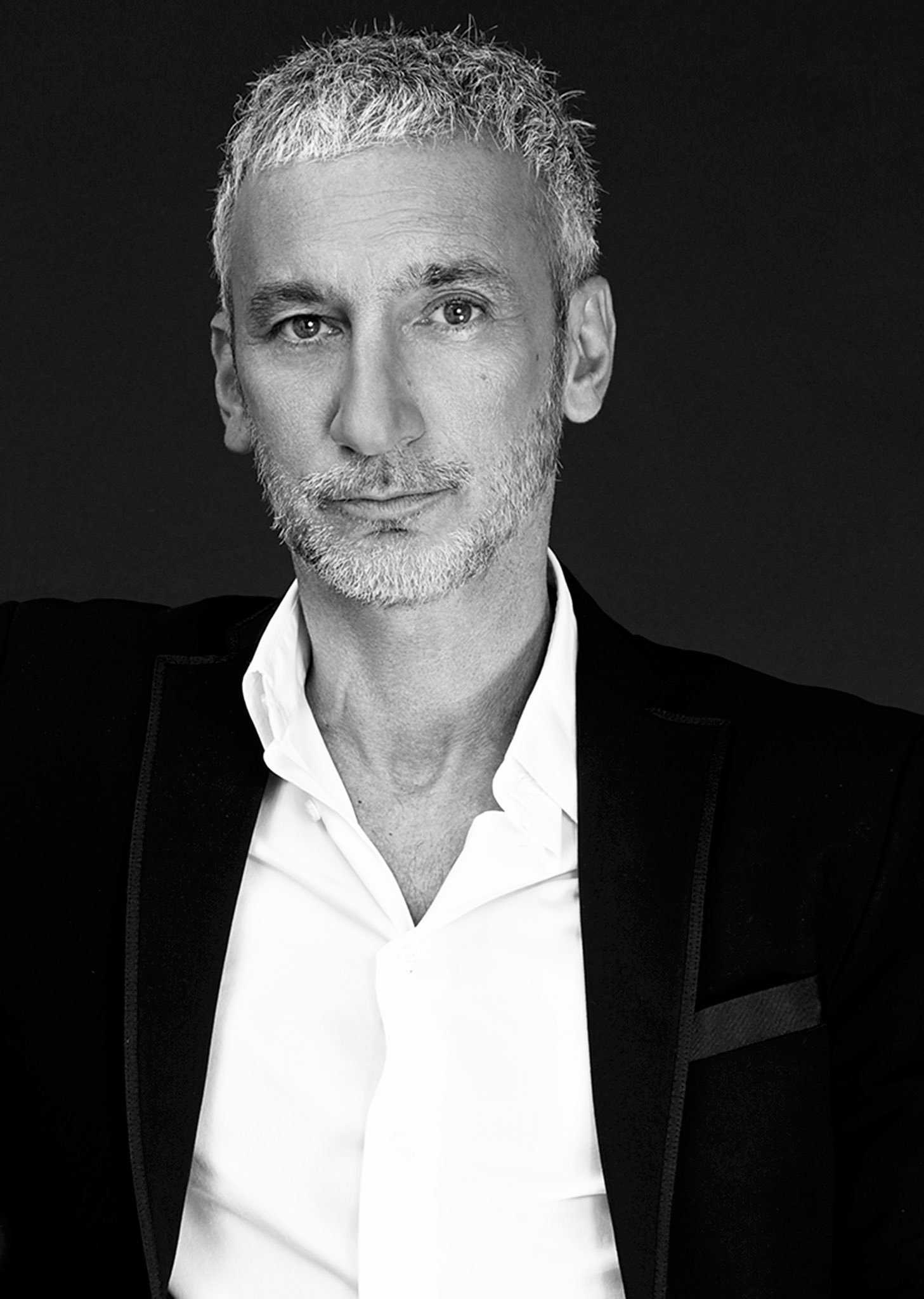 Black-and-white portrait of Akin Konizi with a short, textured silver crop and close-cropped beard, in a black jacket over an open white shirt