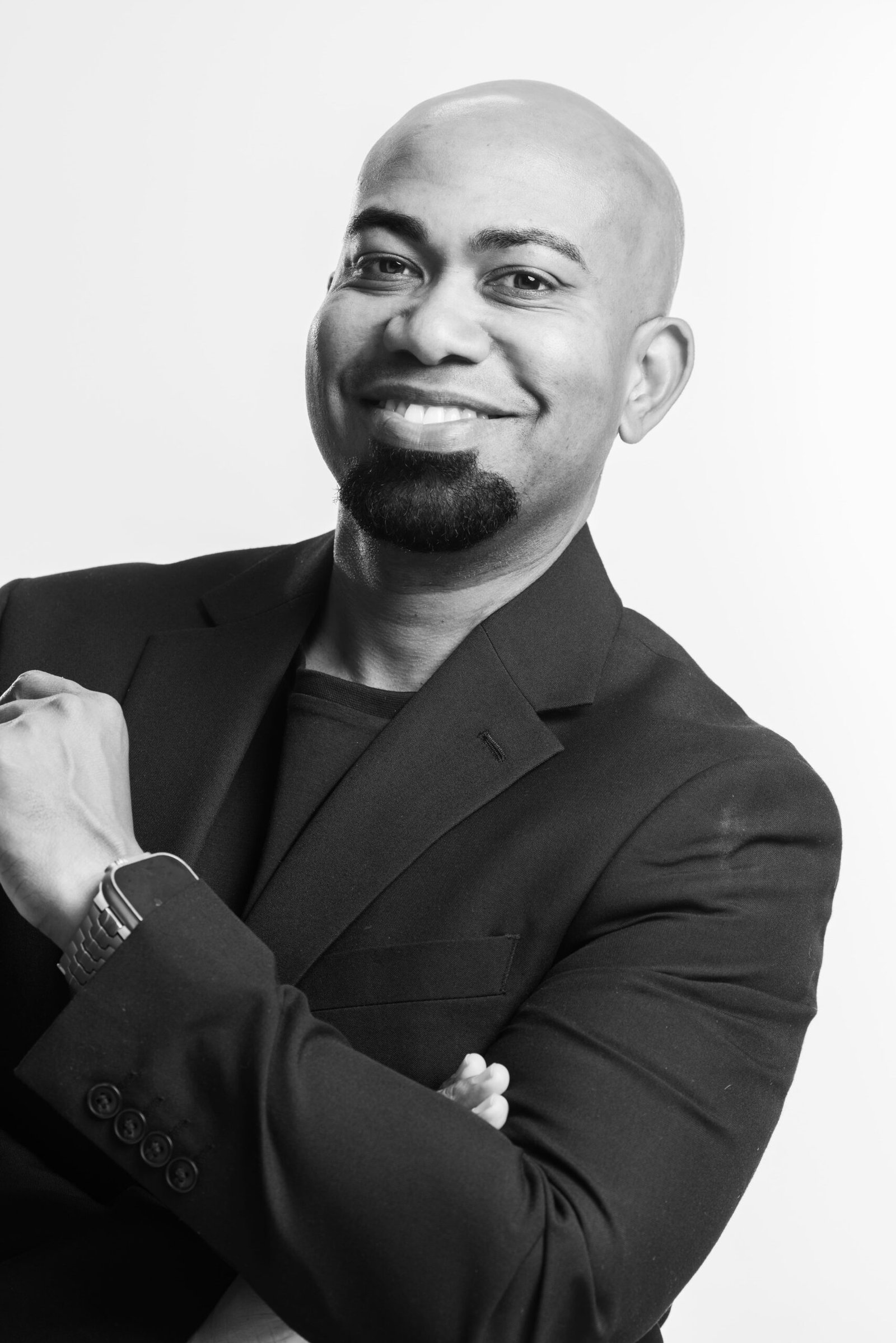 Chris Foster, men's hair specialist and educator, in a black-and-white studio portrait with a closely shaved head and a neatly trimmed goatee, wearing a dark suit jacket over a dark crew-neck, arms folded.
