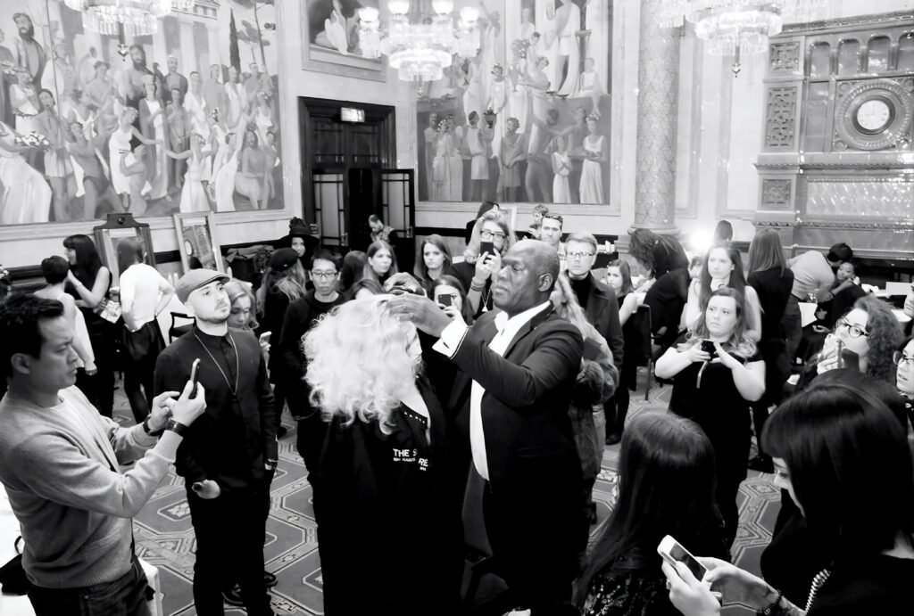 Errol Douglas shapes a model's voluminous, shoulder-length platinum-blonde curls during a live demonstration while hairdressers film on their phones.
