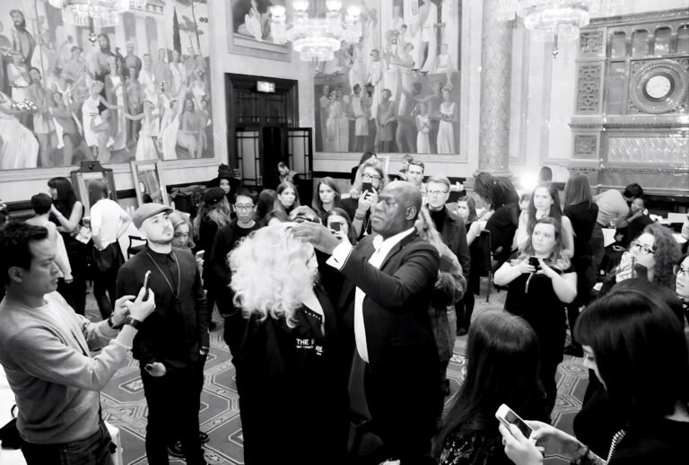 Errol Douglas shapes a model's voluminous, shoulder-length platinum-blonde curls during a live demonstration while hairdressers film on their phones.