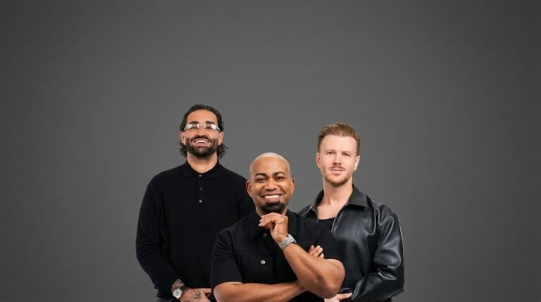 Three male hair professionals in a black-on-black studio group portrait: on the left, dark hair swept back into a low knot with a full trimmed beard and fine silver-rimmed glasses; in the center, Chris Foster with a closely shaved head and a short chin beard, arms folded with one hand raised to his jaw; on the right, light brown hair with tapered sides and volume brushed back from the face, a short mustache and stubble, worn with a black leather jacket.