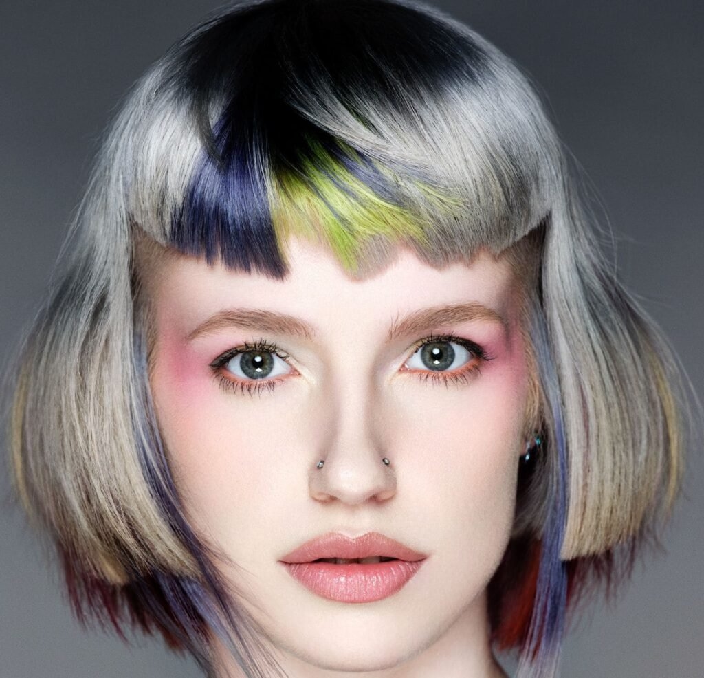 Chin-length silver-blonde bob with a sleek, rounded shape and a high zigzag-edged fringe blocked in black, violet and chartreuse, with fine red and blue strands falling through the ends.