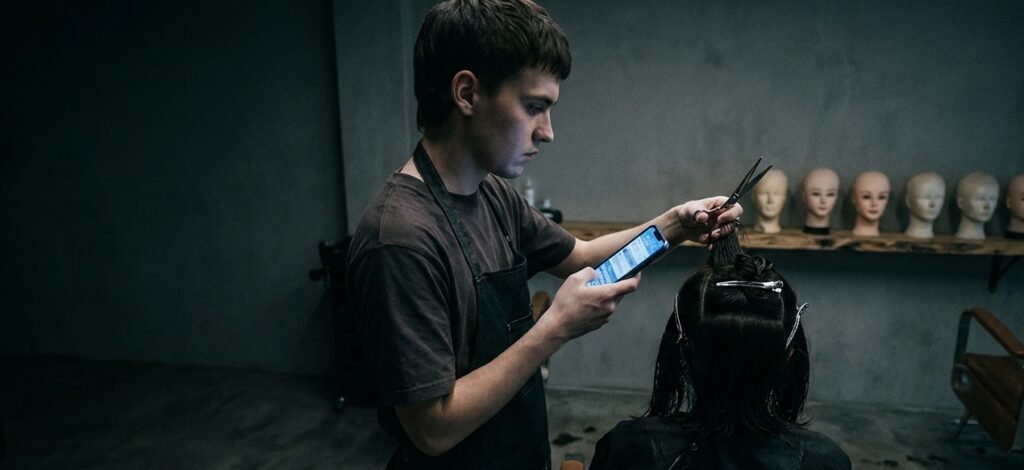 Young hairdresser holding a phone in one hand while lifting a section of a seated client's wet, clipped hair with scissors, training heads on a shelf behind