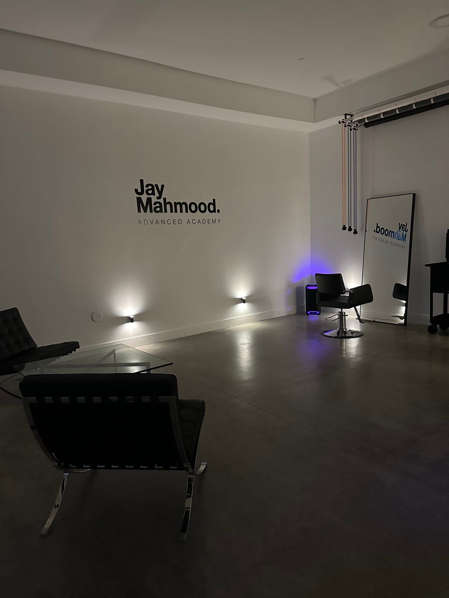 Interior of the Jay Mahmood Advanced Academy with the academy's name on a white wall, a single styling chair, a full-length mirror and black leather lounge chairs.