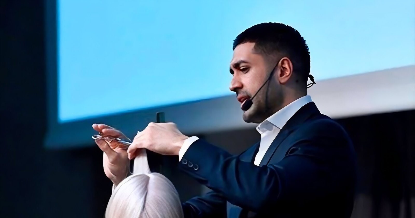 Jay Mahmood lifts a section of icy platinum, shoulder-length straight hair straight up between his fingers while cutting a model's bob with a long side fringe on stage.