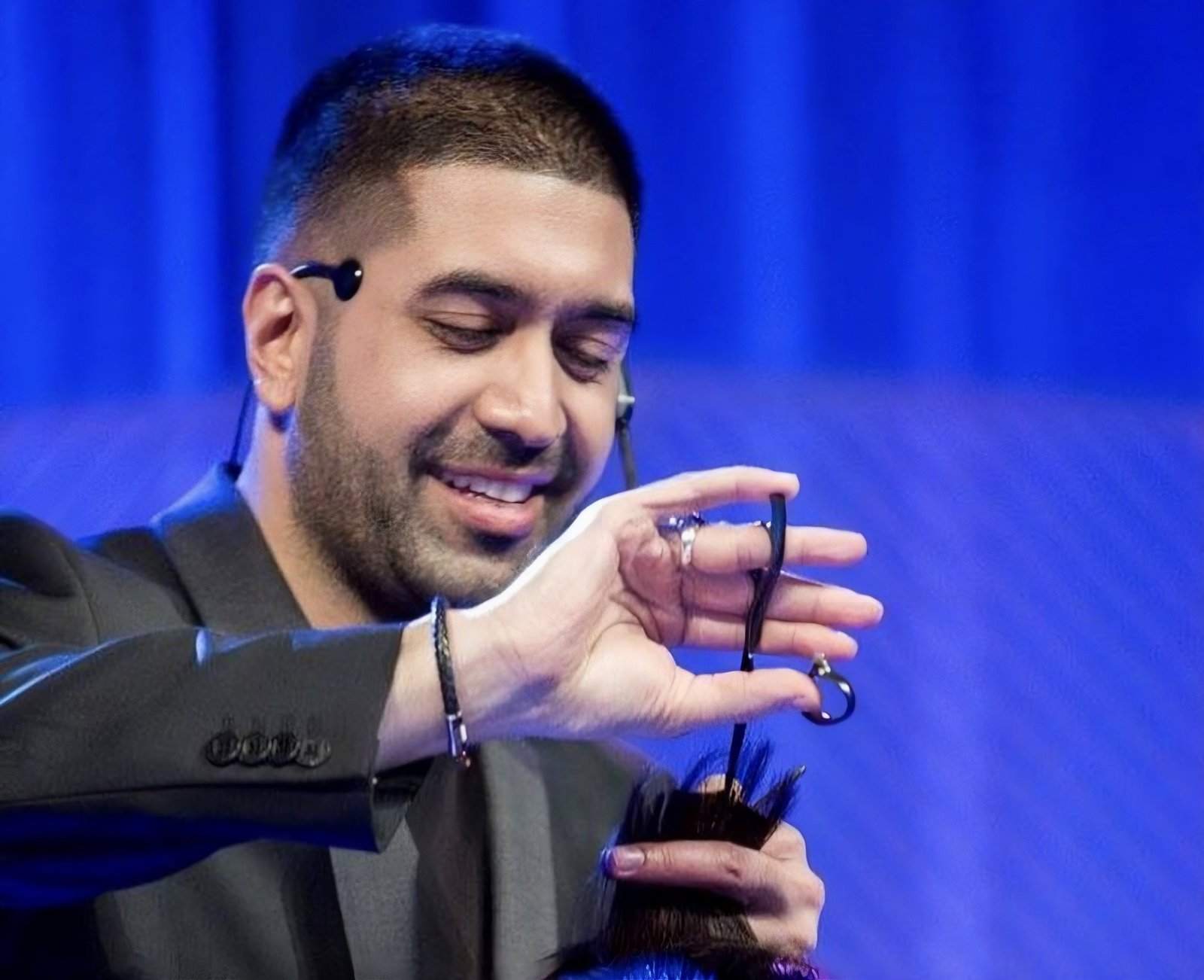 Smiling Jay Mahmood holds scissors open in his palm while working through a section of dark hair during a live demonstration.
